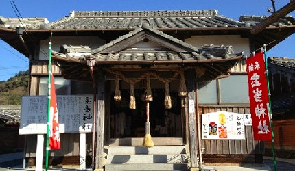 木造日式神社本殿入口,棕色瓦屋顶下悬挂大麻绳和铃铛,左右插有红色旗帜,上书『宝当神社』字样