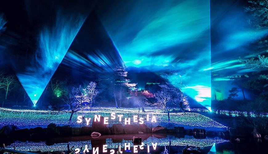 夜空下的山林被藍綠色雷射與霧光包圍,「SYNESTHESIA」字樣在水面上映出光影倒影