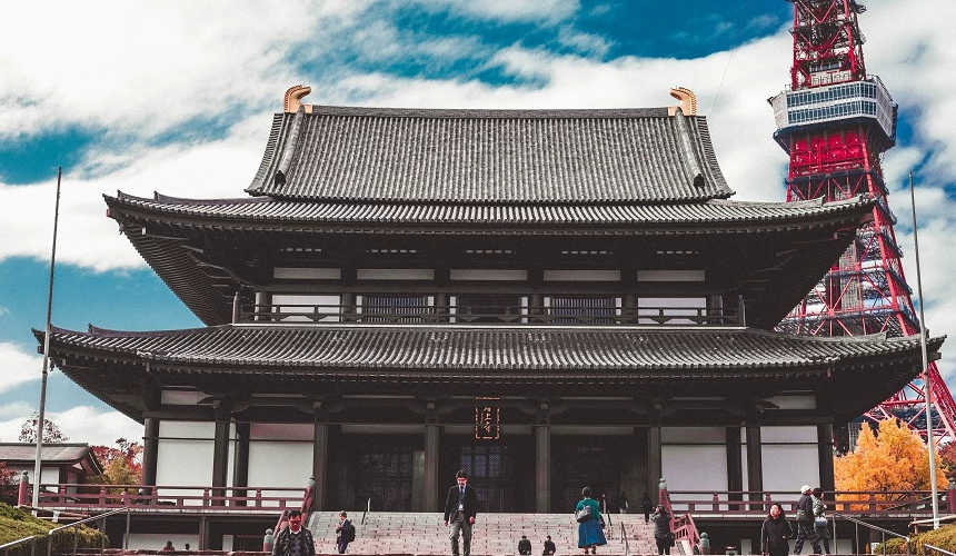 正面拍攝的增上寺本堂,灰色屋頂氣勢宏偉,背景右側可見紅色東京鐵塔,遊客散步在石階與廣場上