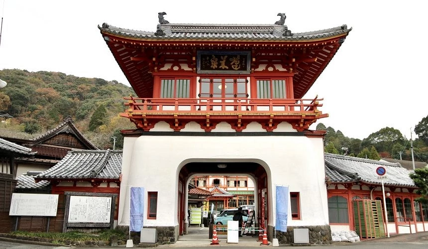 武雄温泉入口大门,白色拱形下层与红色二层楼阁式屋顶,门洞内可见停放的车辆与延伸至后方的温泉建筑,背景为绿意山坡。