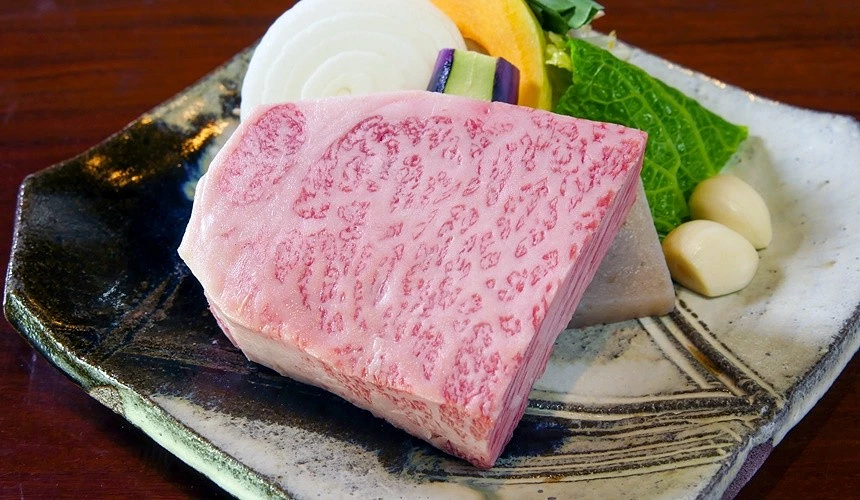一块油花浓郁的厚切佐贺牛生肉,旁边摆有洋葱切片、南瓜片、茄子、绿叶蔬菜和蒜瓣,盛放在日式陶瓷盘上。