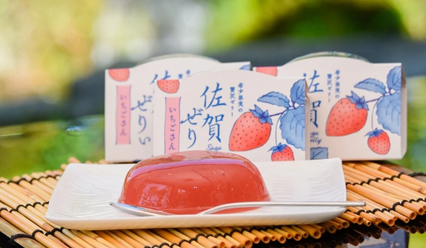 白色方盘上摆放着透亮鲜红的草莓果冻,旁边放着银色小汤匙,后方则排列着印有草莓插画与日文「佐贺ゼリー」字样的包装盒。