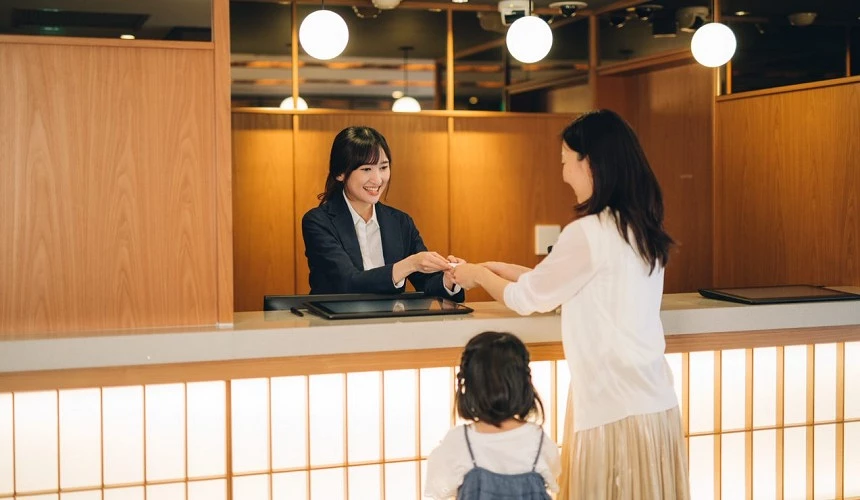 MIMARU 飯店櫃檯人員面帶微笑接待一位女性與小孩,櫃台設計以木質調為主,展現親切且溫馨的日式服務氛圍