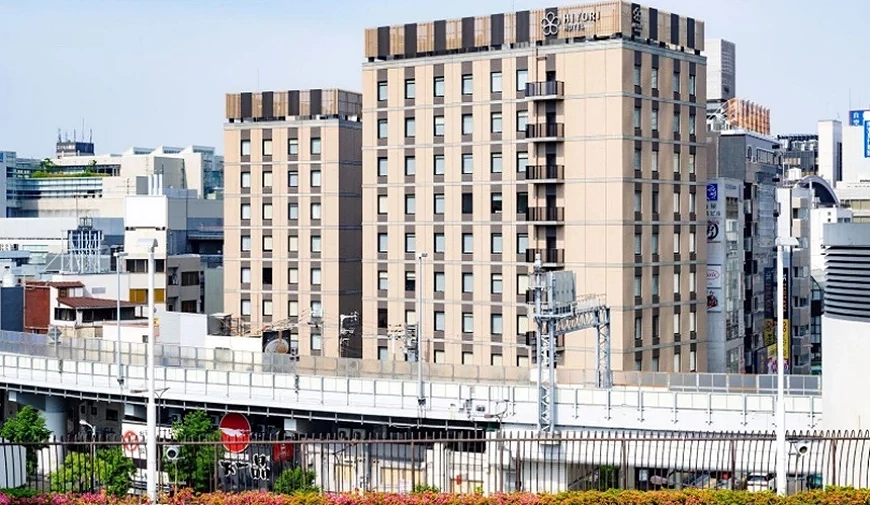 两栋米色高楼并列的饭店建筑,顶楼挂有 HIYORI HOTELS 招牌,前景为高架道路与铁道轨道,背景可见都市天际线。