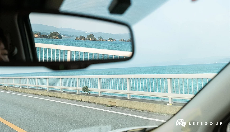 从车内视角拍摄冲绳海岸公路,窗外为蓝色海洋与护栏,后照镜中映出远方岛屿景色