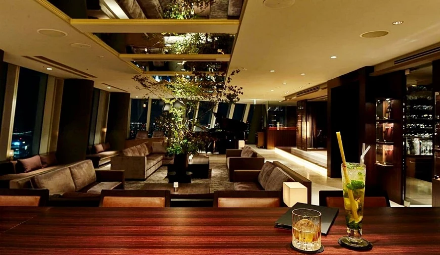高樓層夜景酒吧沙發區,木質調空間搭配舒適沙發與綠意植栽,桌上擺有雞尾酒與威士忌,營造靜謐雅緻的休憩氛圍。