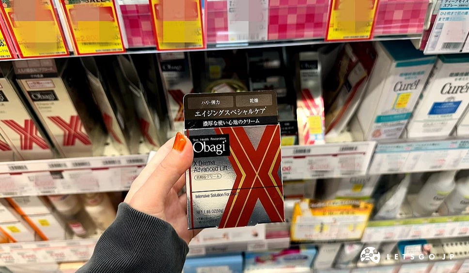 一位女性的手拿着一盒上面印有英文Obagi字样及一个大大红色英文字母和黄色线条的X字的银色正方形纸盒站在商品架前