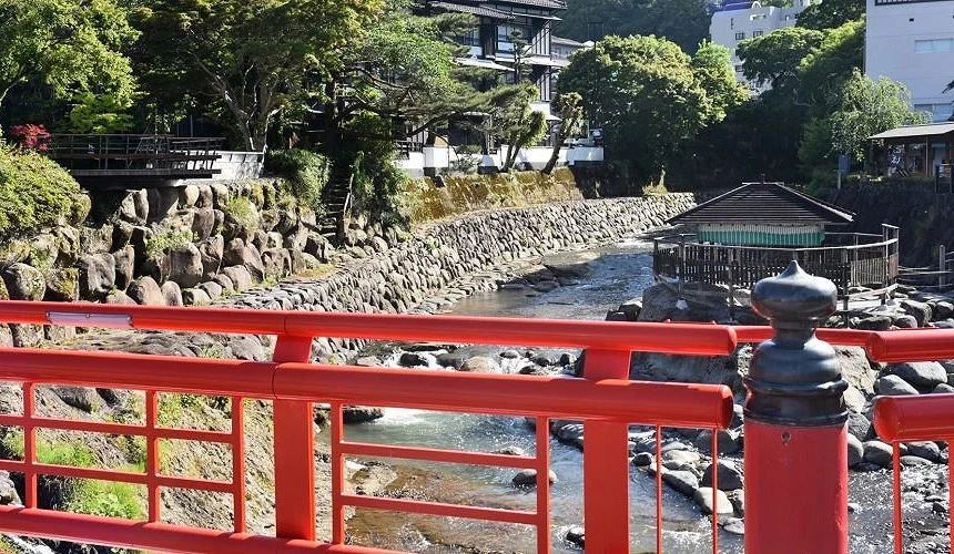 伊豆修善寺温泉街的红色栏杆桥与清澈溪流,背景是传统日式建筑与绿意盎然的自然景观。