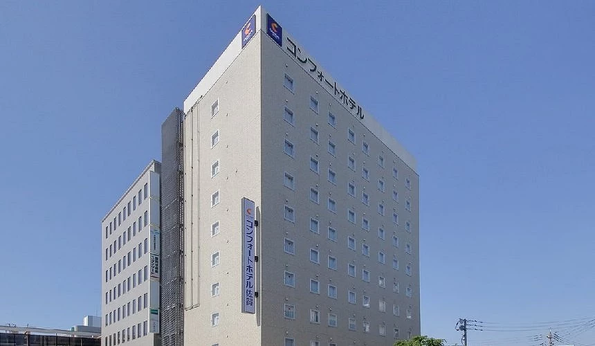 蓝天映衬下的Comfort Hotel佐贺大楼,米色砖墙外观上整齐排列多扇小窗,顶楼与立面淡蓝色招牌标示饭店名称