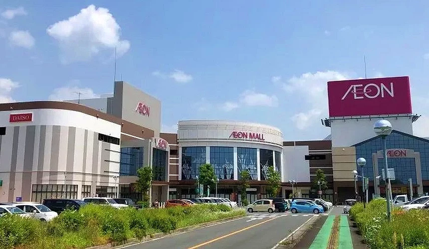 蓝天白云下,一条道路两旁停满汽车,尽头为大型AEON购物中心,中央圆弧玻璃立面,右侧高挂AEON红色招牌,左侧可见DAISO店招