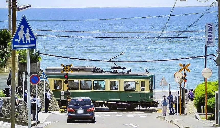 後面是一大片海,前面有一個綠黃色火車通過,有幾個行人和一台車在平交道前
