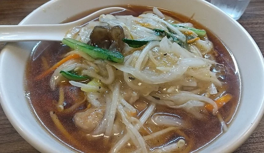 白色碗裡裝有湯麵,上面有綠色蔬菜和橘色紅蘿蔔絲,左上角有湯匙叉在碗裡