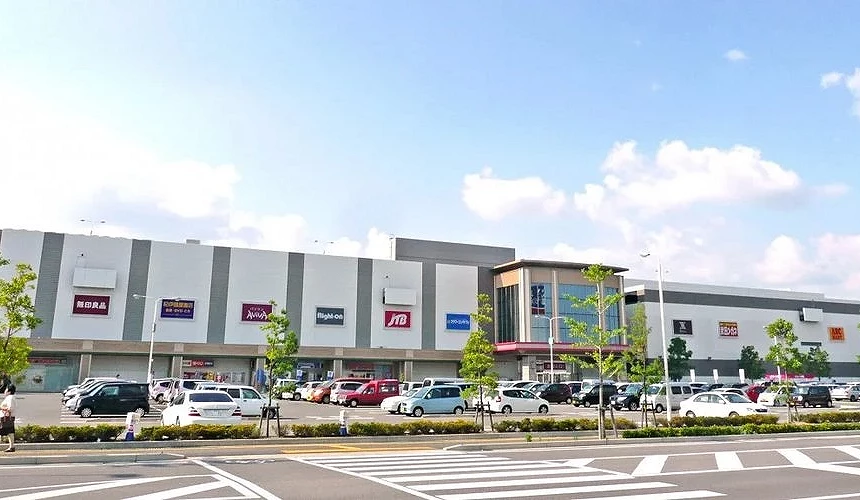 蓝天白云下的购物中心外观,立面上有无印良品、JTB、Right-on等店铺招牌,前方停车场停满汽车,路旁有人行道