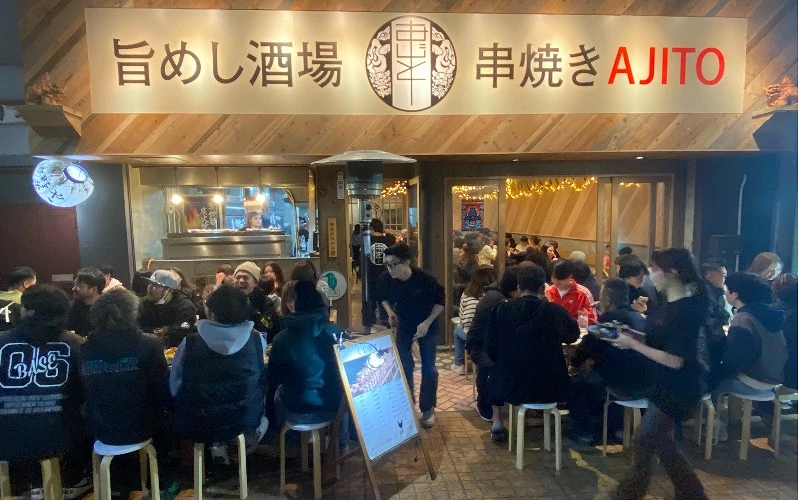 串燒居酒屋外觀，門口與室內皆坐滿客人，招牌寫有「旨めし酒場 串焼き AJITO」