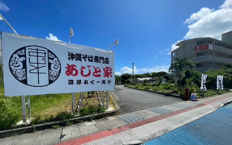 藍天下道路旁立牌，上寫「沖繩そば専門店 あじと家」