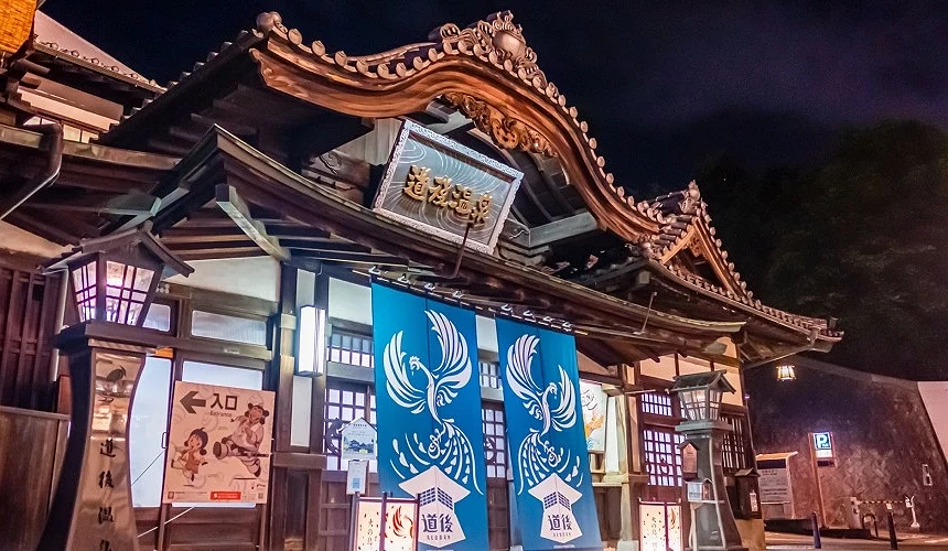 道后温泉本馆夜间景观,传统木造建筑配上蓝色暖帘与凤凰图腾