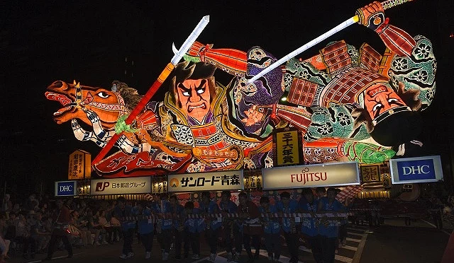 在夜晚的祭典上,一座巨大的彩色花燈展示著武士揮劍戰鬥的場景,由許多人抬著在街道上前進。