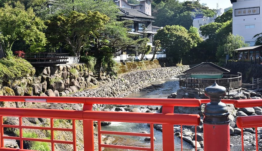 修善寺温泉街的红色栏杆桥与河畔「独钴之汤」温泉设施，背景为传统旅馆与绿意盎然山林景观