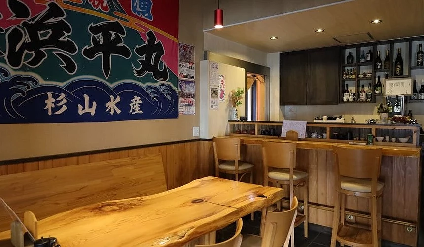 杉山水产 Sugiya 店内用餐空间,木制吧台与长桌搭配墙面大型渔旗与日本酒展示,营造出温馨朴实的日式氛围