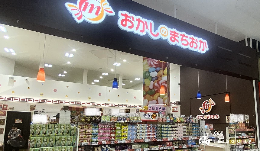 零食小舖店鋪入口,上方有日文招牌「おかしのまちおか」,內部貨架上陳列著豐富的零食。