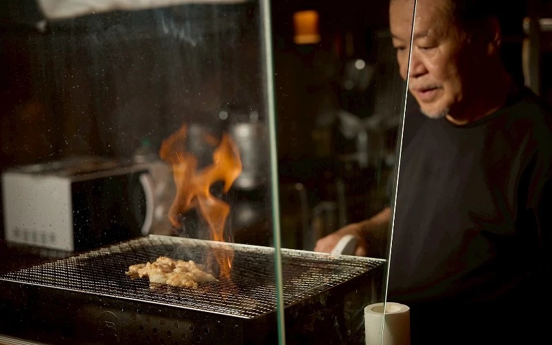 廚師在烤爐上以火焰燒烤肉塊，前方有玻璃隔板
