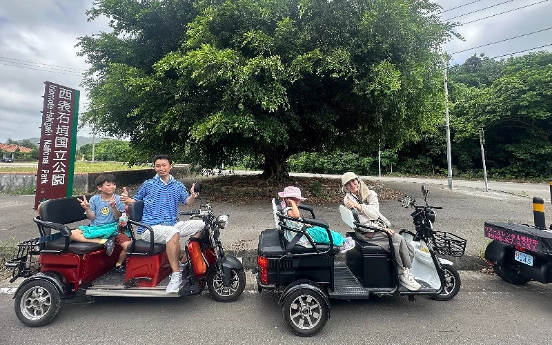 兩台三輪車分別載著兩人，停駐於一棵大樹前