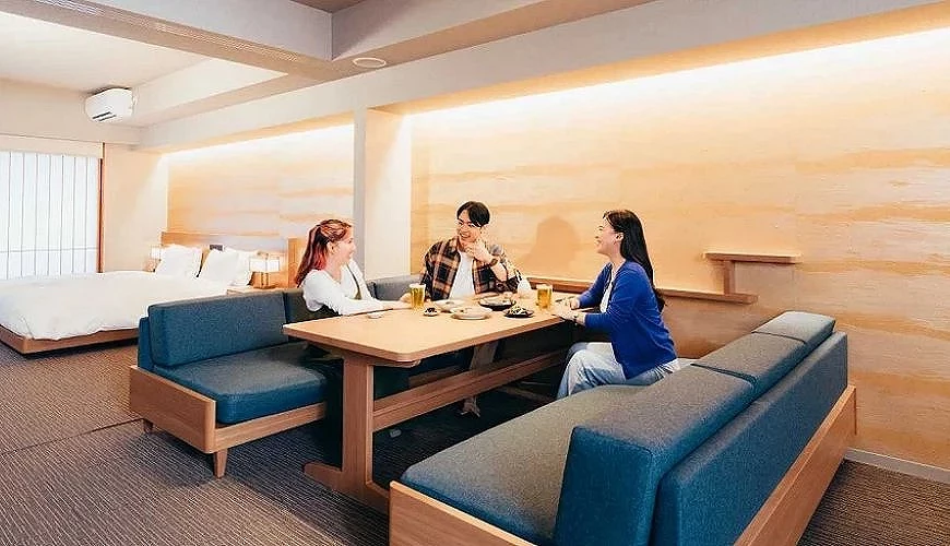 三位旅客在「APARTMENT HOTEL MIMARU 大阪」寬敞的客房中享用餐點,房內設有餐桌、沙發與雙人床,呈現舒適的公寓式住宿空間