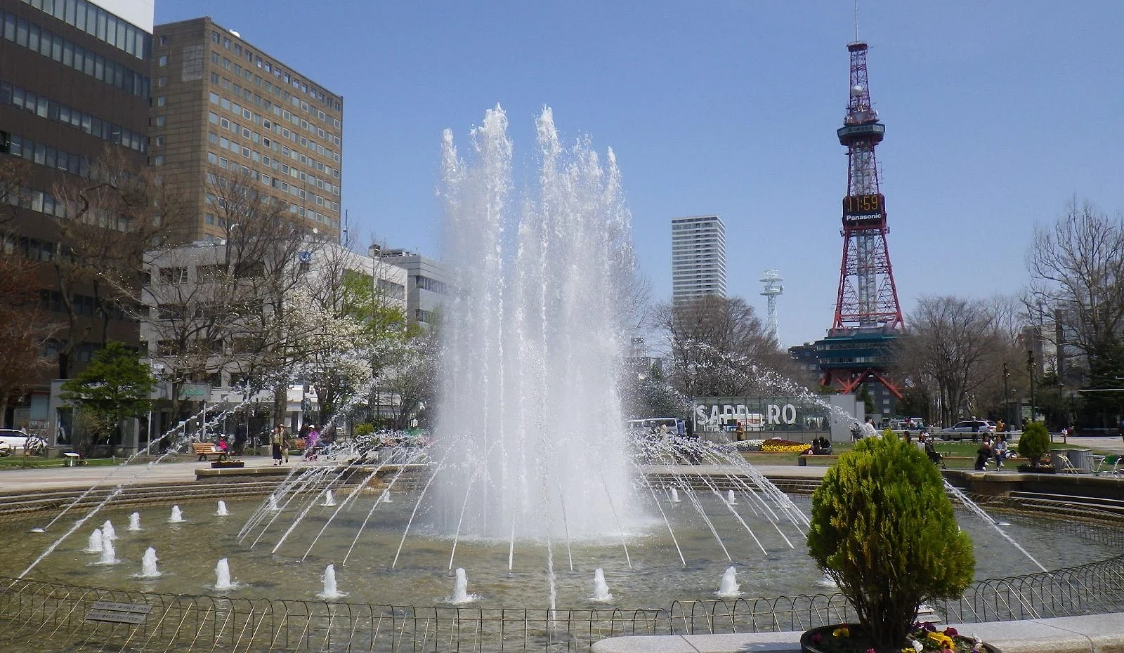 札幌大通公园喷泉广场景象,中央水柱高高喷起,背景为红白相间的札幌电视塔与蓝天