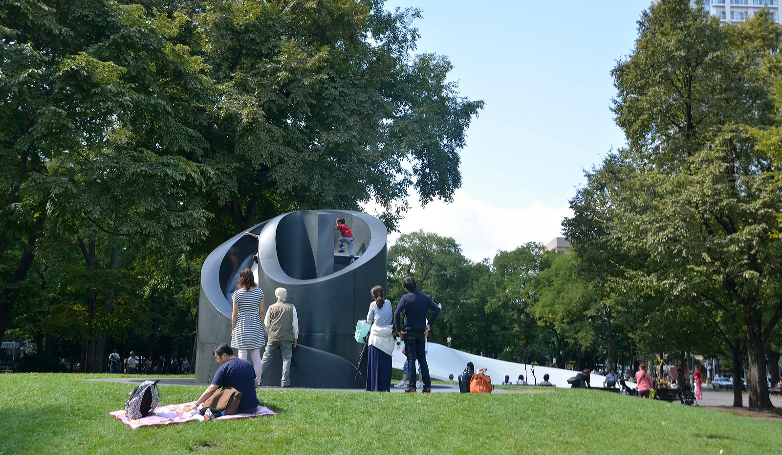 札幌大通公园中,一座黑色螺旋形的艺术溜滑梯吸引许多民众驻足观看与游玩,前景有人坐在草地上野餐,背景是高大的绿树与现代建筑