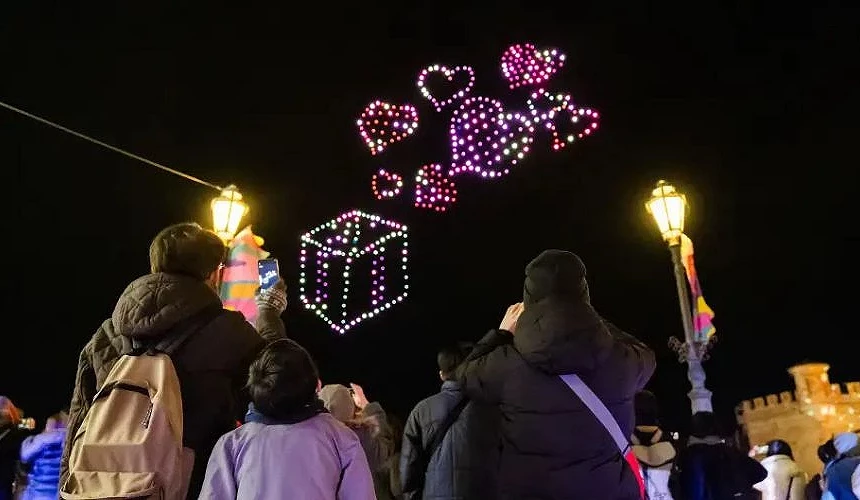 觀眾抬頭欣賞夜空中無人機組成的禮物盒與愛心圖案燈光秀,街燈與人群映襯出節慶熱鬧氛圍