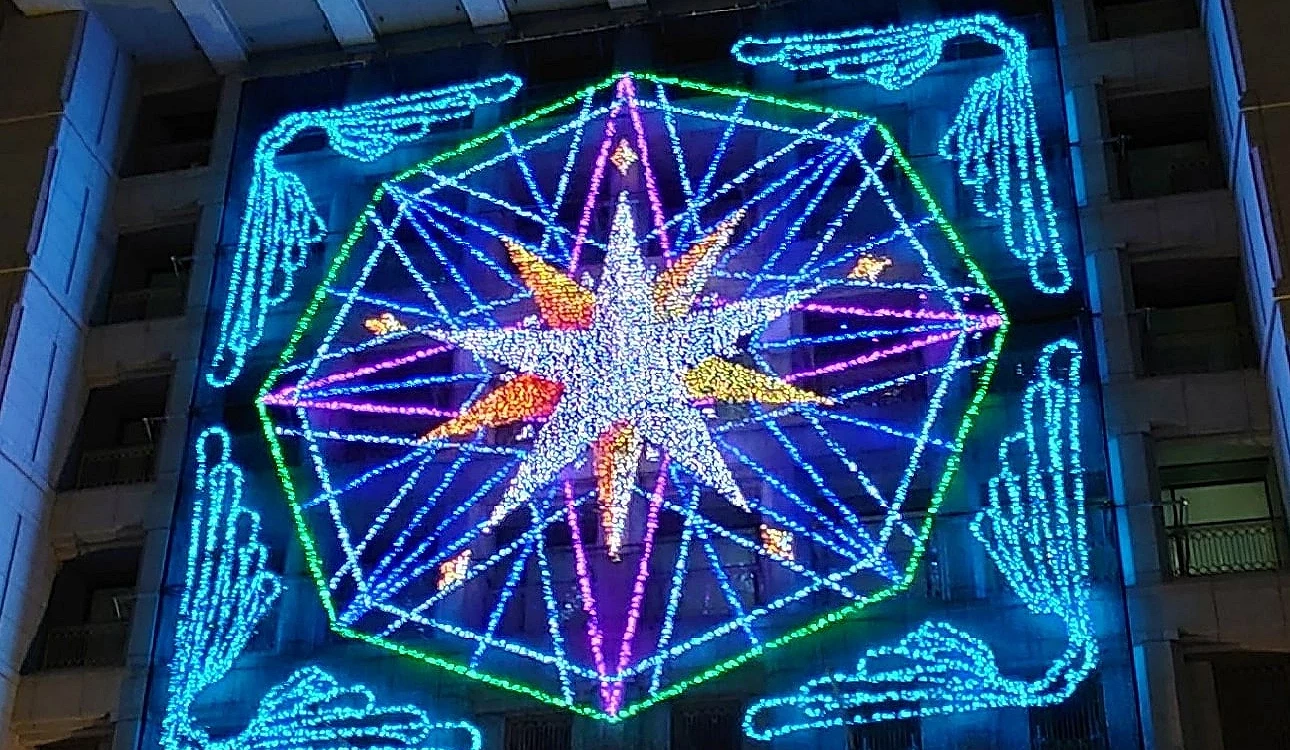 夜間建築外牆上以燈飾拼成巨大的八角星圖案,中心閃耀著橙色與藍紫色光芒,周圍線條構成幾何放射狀設計