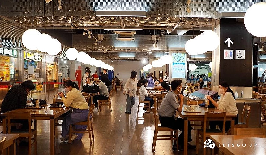 成田机场第三航厦2楼的美食广场,旅客坐在木质桌椅区用餐,后方可见各式餐厅与明亮的灯饰,气氛热闹舒适