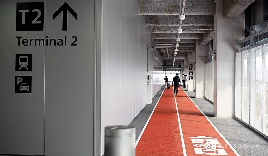 成田机场第三航厦与第二航厦之间的连接通道,红色地面导引线标示「Terminal 2」方向,旅客沿着指示步行前往第二航厦