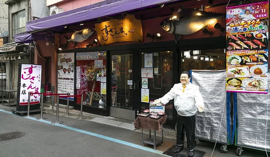 「壽司三味本店」外觀,入口上方掛著大型鮪魚模型與木製店牌,店門前擺有創辦人雕像與壽司展示模型
