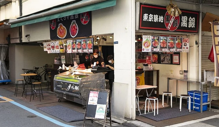 「まぐろや黒銀 築地本店」外觀,黑白招牌上印有鮪魚圖案與店名,店門前擺放新鮮魚貨展示冰台與菜單看板,內部可見店員忙碌製作壽司與海鮮丼