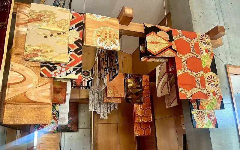 多條帶有金色與紅色圖紋的和服腰帶懸掛於木架上