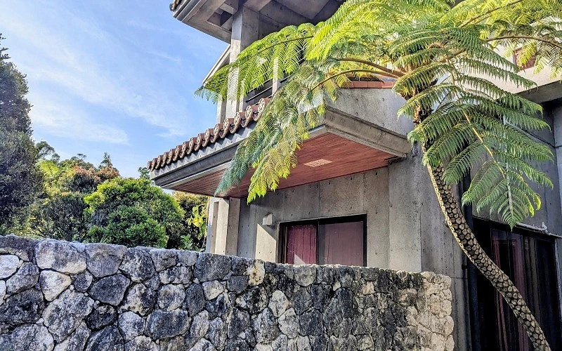 灰色石牆與混凝土建築外觀，屋簷下有紅瓦與樹蕨