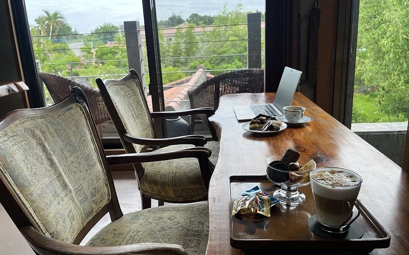 木桌上擺放筆電、咖啡與點心，窗外可見綠樹與屋頂景色
