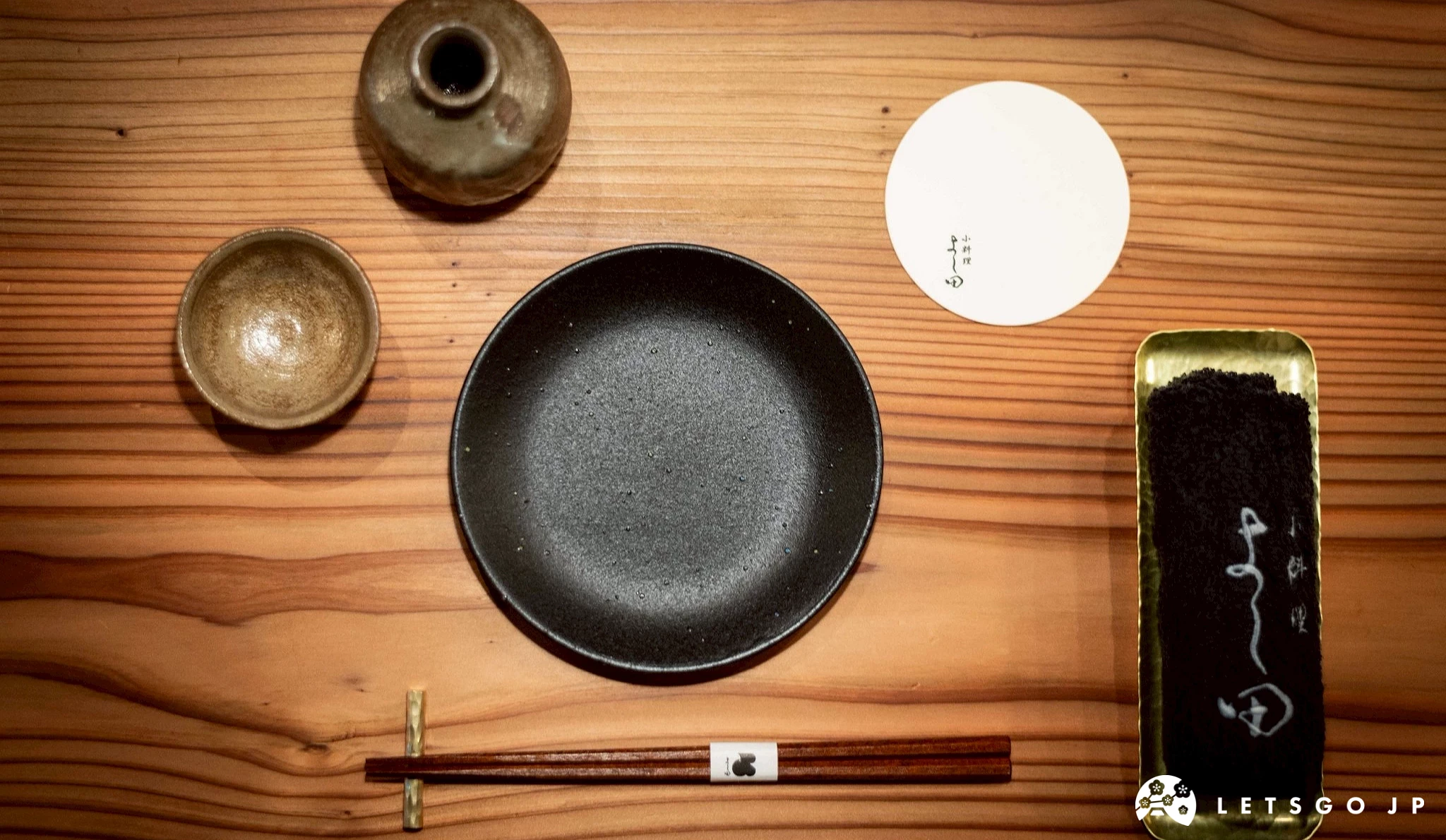 俯視角度拍攝的日式餐桌擺設,包括一個黑色的陶製圓盤、筷子、小酒杯和杯墊,展現用餐儀式感。