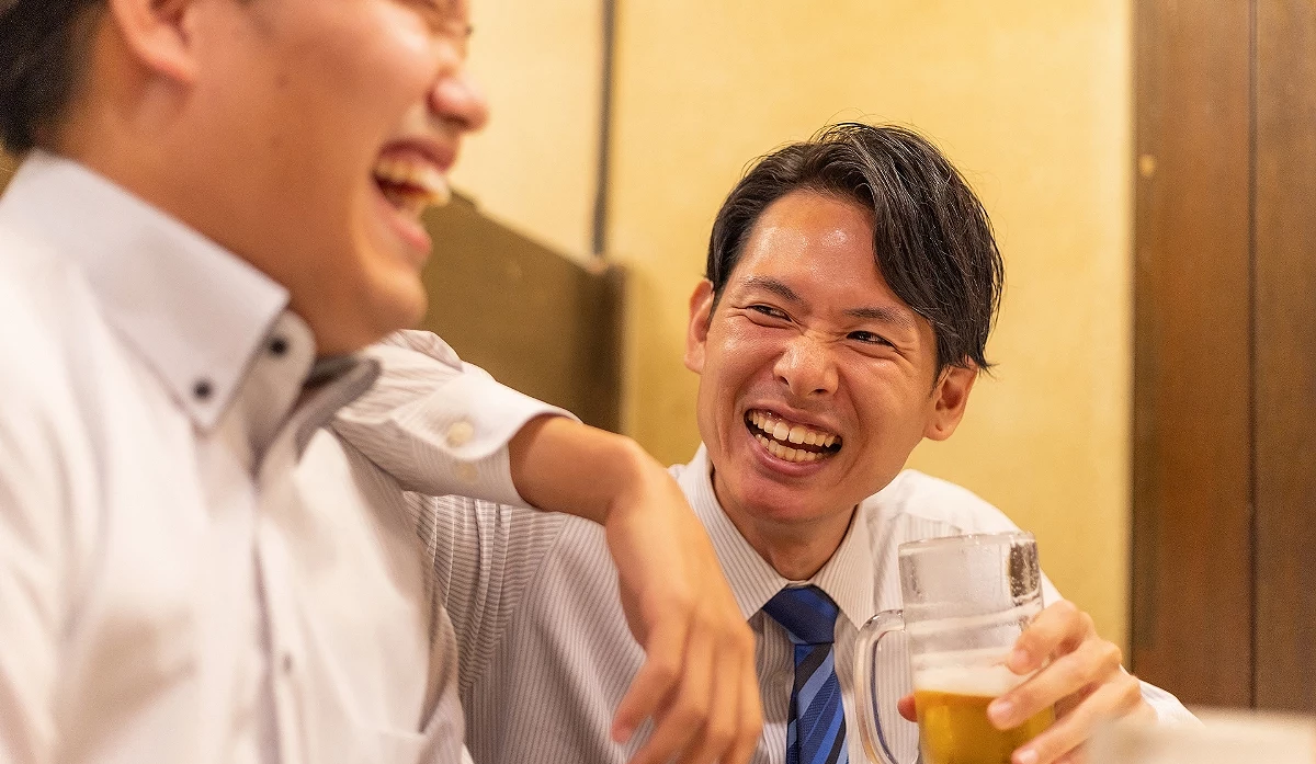 兩位穿著西裝的日本男性上班族在居酒屋內,其中一位舉著酒杯,熱情地大笑交談。
