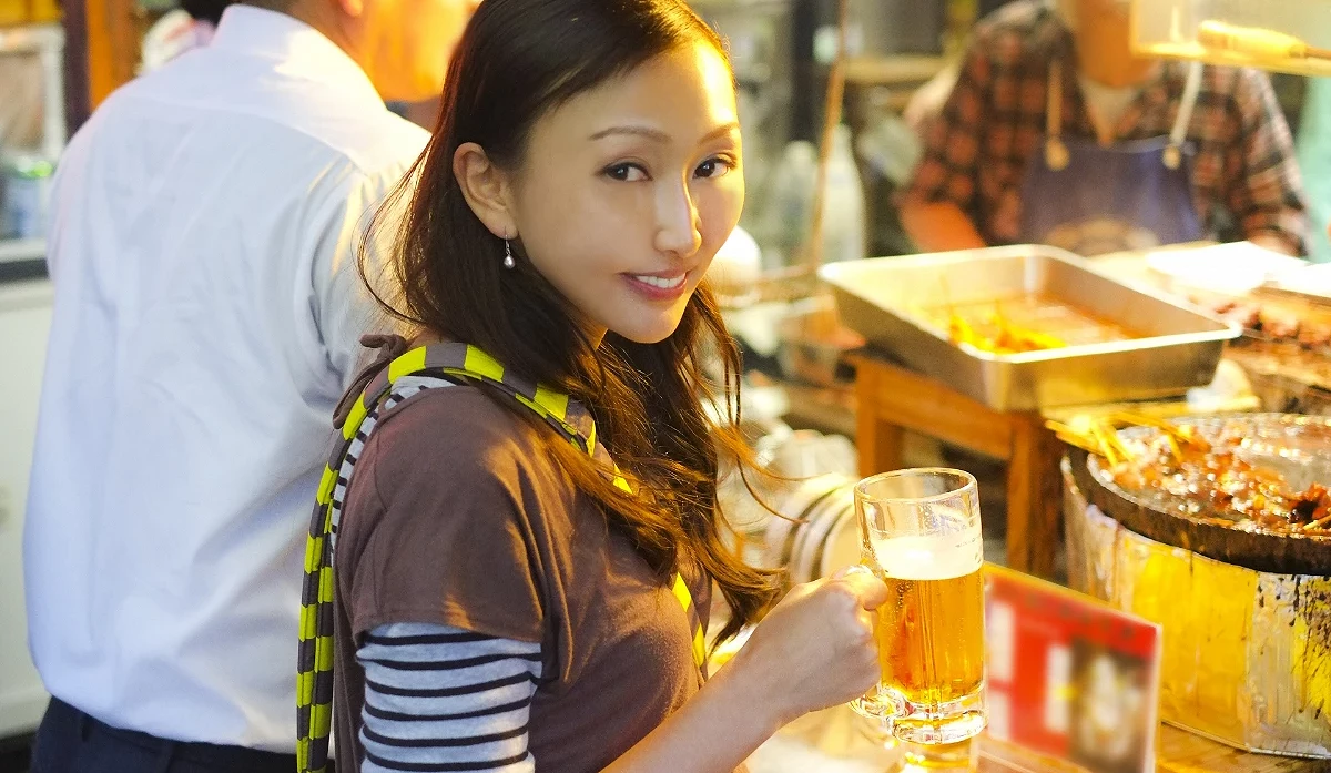 位穿著黃色條紋上衣的年輕女性,在居酒屋的暖黃燈光下拿著一杯啤酒微笑。