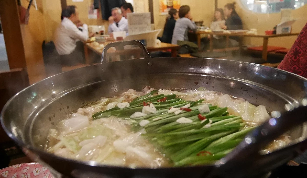 一個裝滿肉丸子和蔬菜的日式火鍋或雜燴正在爐上沸騰,散發熱氣。背景是居酒屋的客人