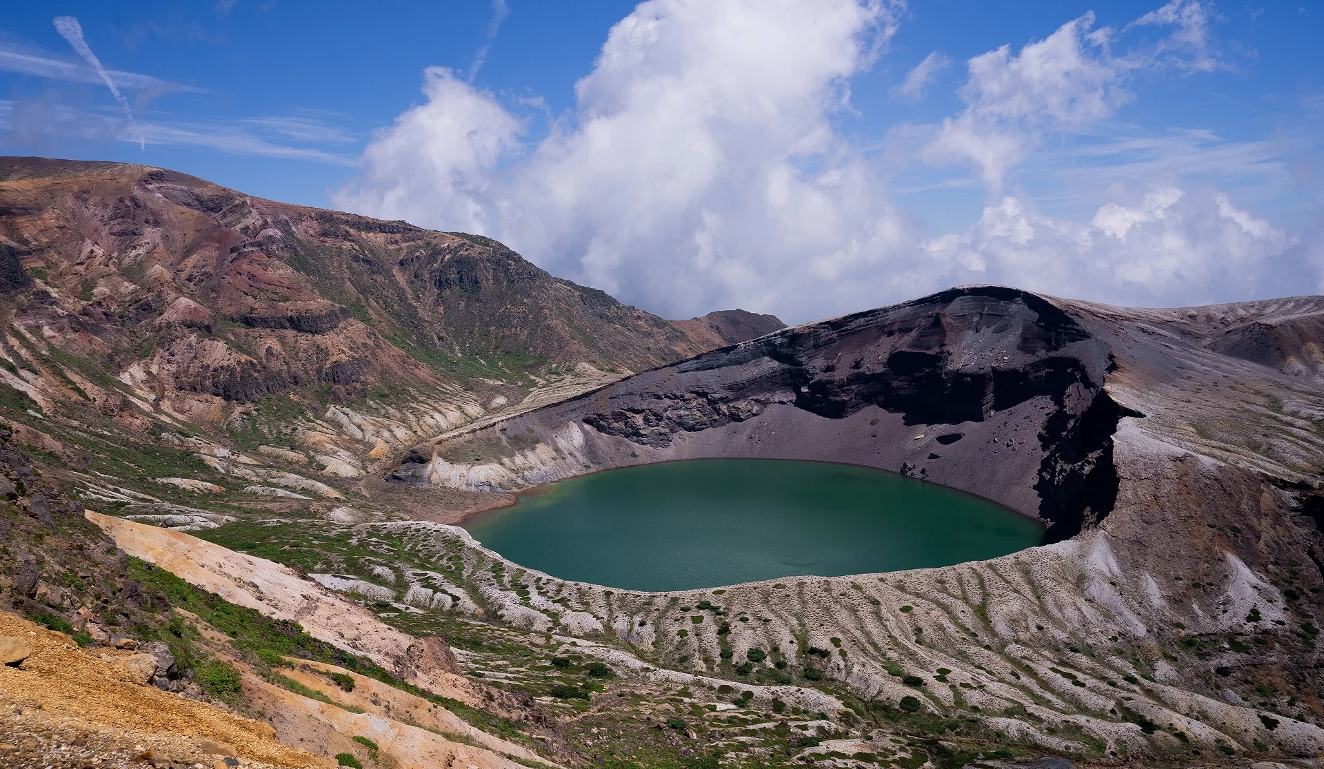 山區火山口中央有碧綠湖水,周圍岩壁層理分明,天空雲層低垂