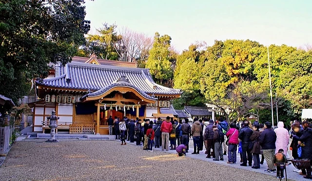 一座傳統日式神社的主殿前,民眾排成長長隊伍等候參拜,四周被樹木與自然景色環繞