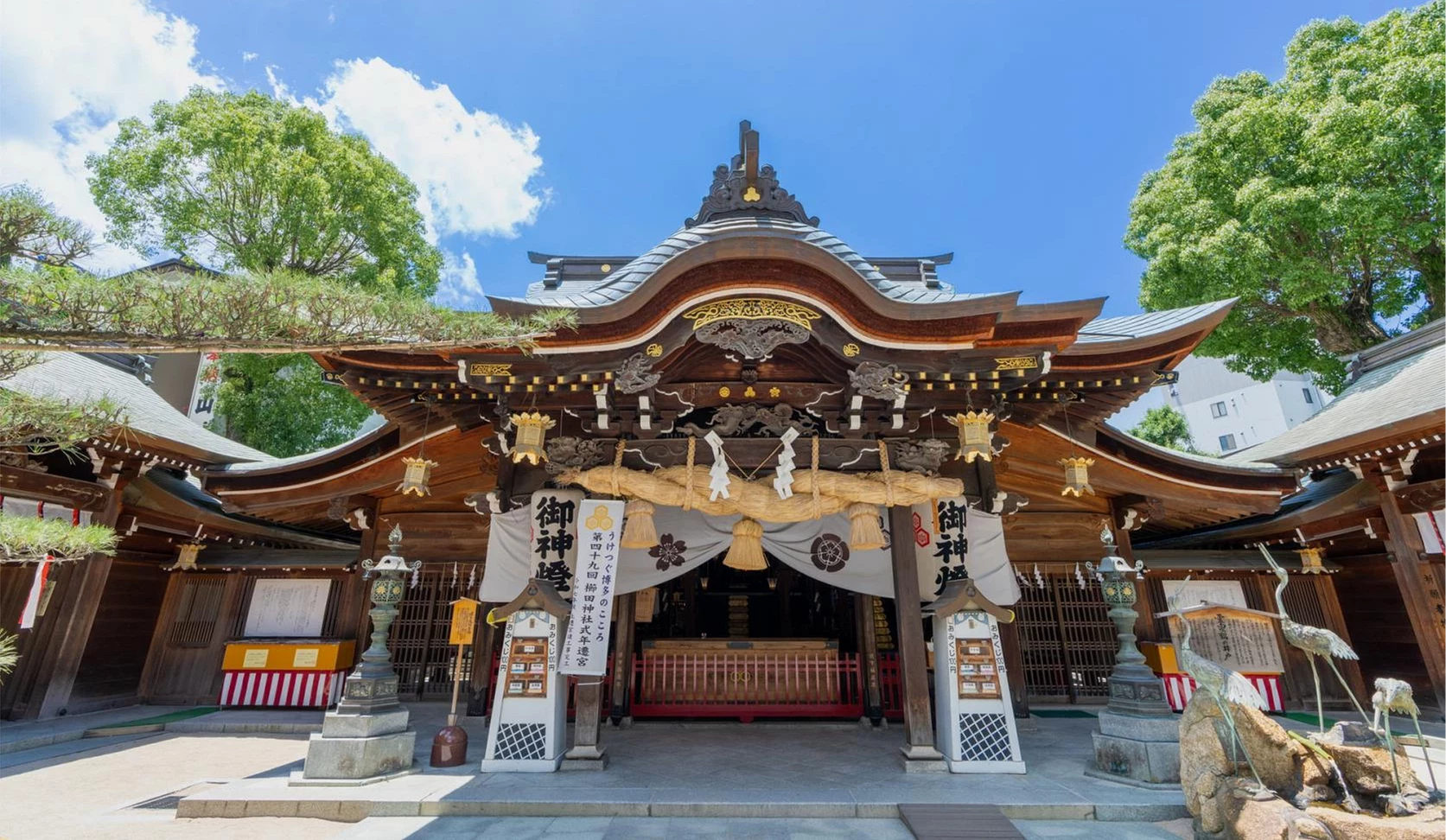 櫛田神社拜殿正面,深色木造屋頂線條優雅,中央掛著大型注連繩,周圍可見樹木