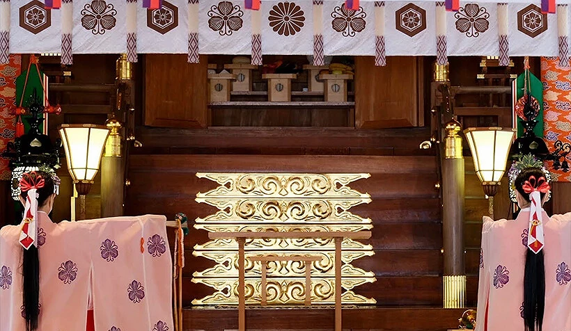 十日惠比須神社神殿內部,可見兩位巫女身穿粉色神樂舞服飾,在金色裝飾前進行儀式,周圍點綴燈光與傳統布幔