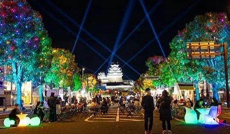 姬路城前圣诞节夜景,街道两侧树木点亮多彩灯饰,天空中有蓝色光束照向城堡,街中央设有摊位与座椅,人潮热闹