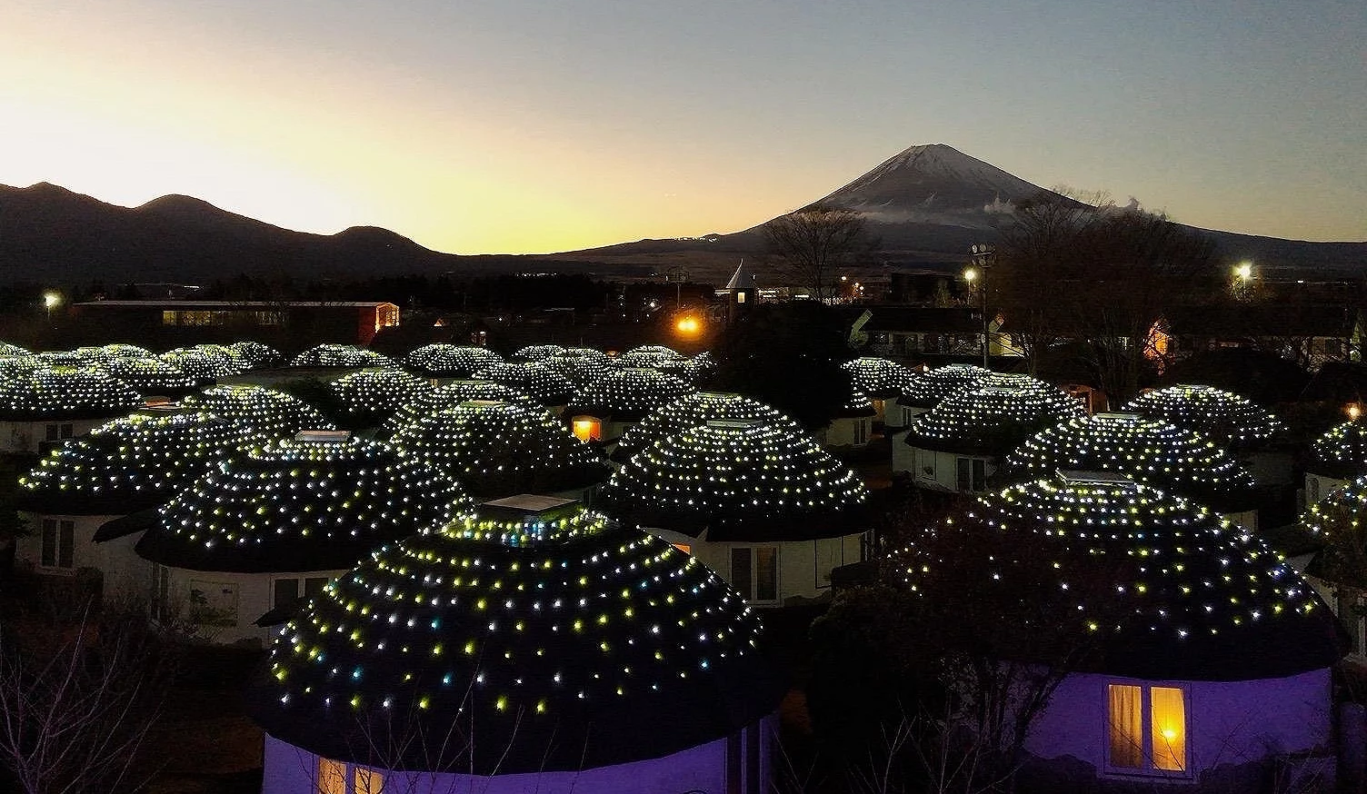 富士山夕景下的御殿场高原时之栖度假村,圆顶小屋屋顶覆满灯饰,点点光芒在夜色中闪烁,远方可见被夕阳染红的富士山与群山轮廓