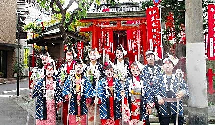 身着和服与法被、脸上化有狐狸妆容的参与者站在王子稻荷神社前,周围可见成排的红色旗帜