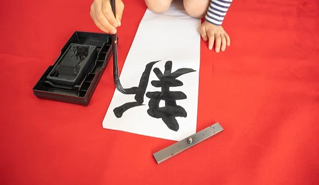 孩童在红色垫布上以毛笔书写新年开笔作品,白纸上写有大大的黑色汉字,呈现日本新年开笔的传统场景